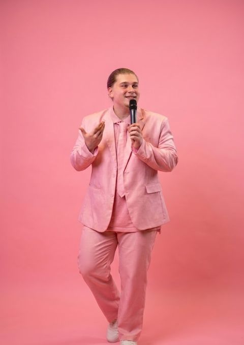 Tom EGE en costume rose complet, micro à la main, en train de parler, sur un fond rose uni.